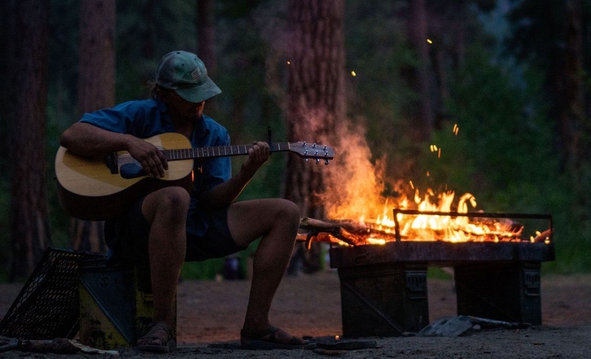 A man is playing a guitar in front of a campfire.