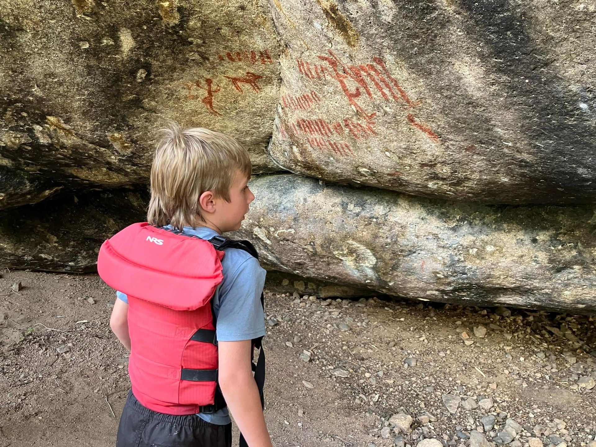 A young boy wearing a life jacket is standing in front of a large rock.