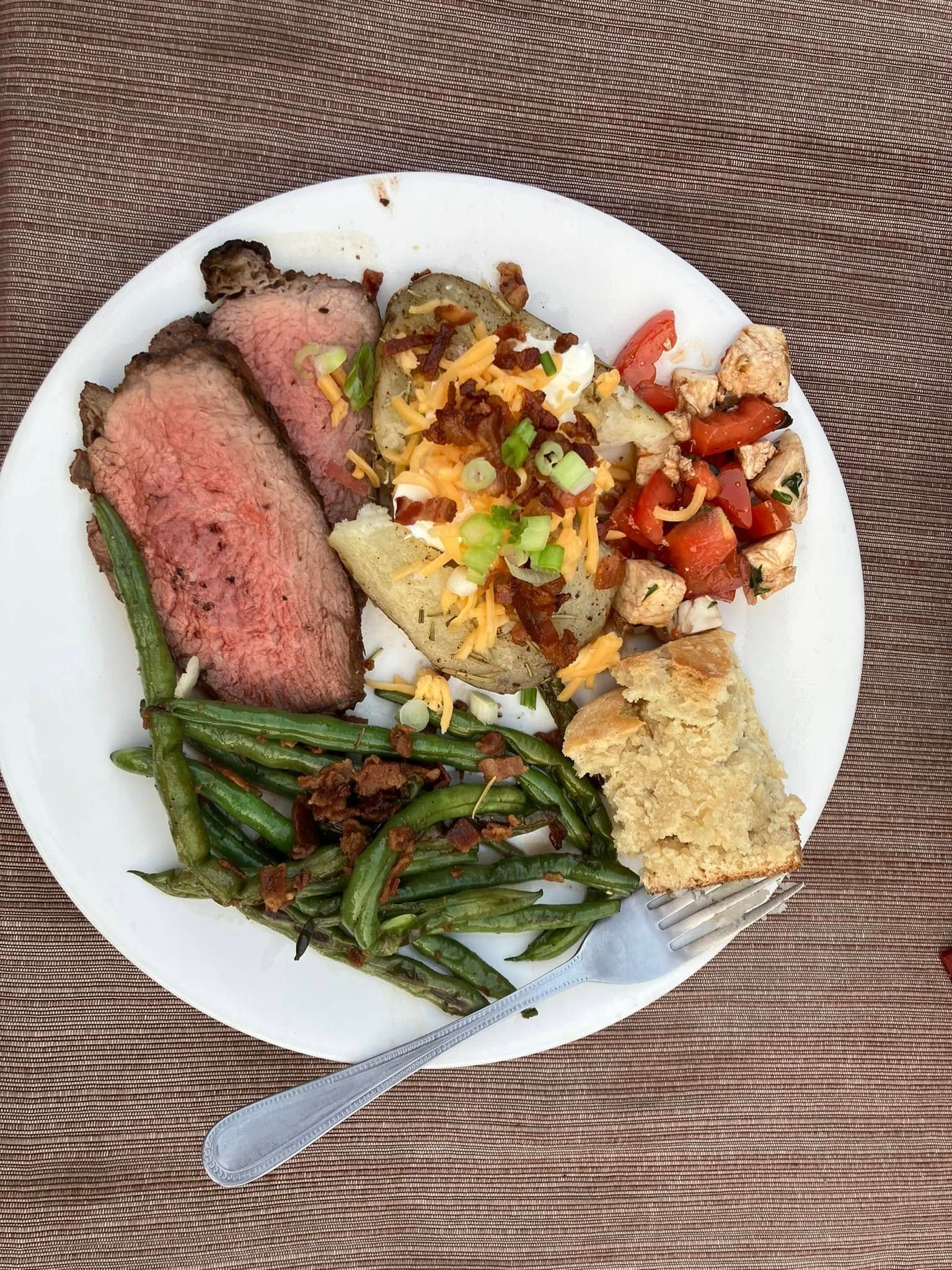 A white plate topped with meat , green beans , tomatoes , cornbread and a fork.