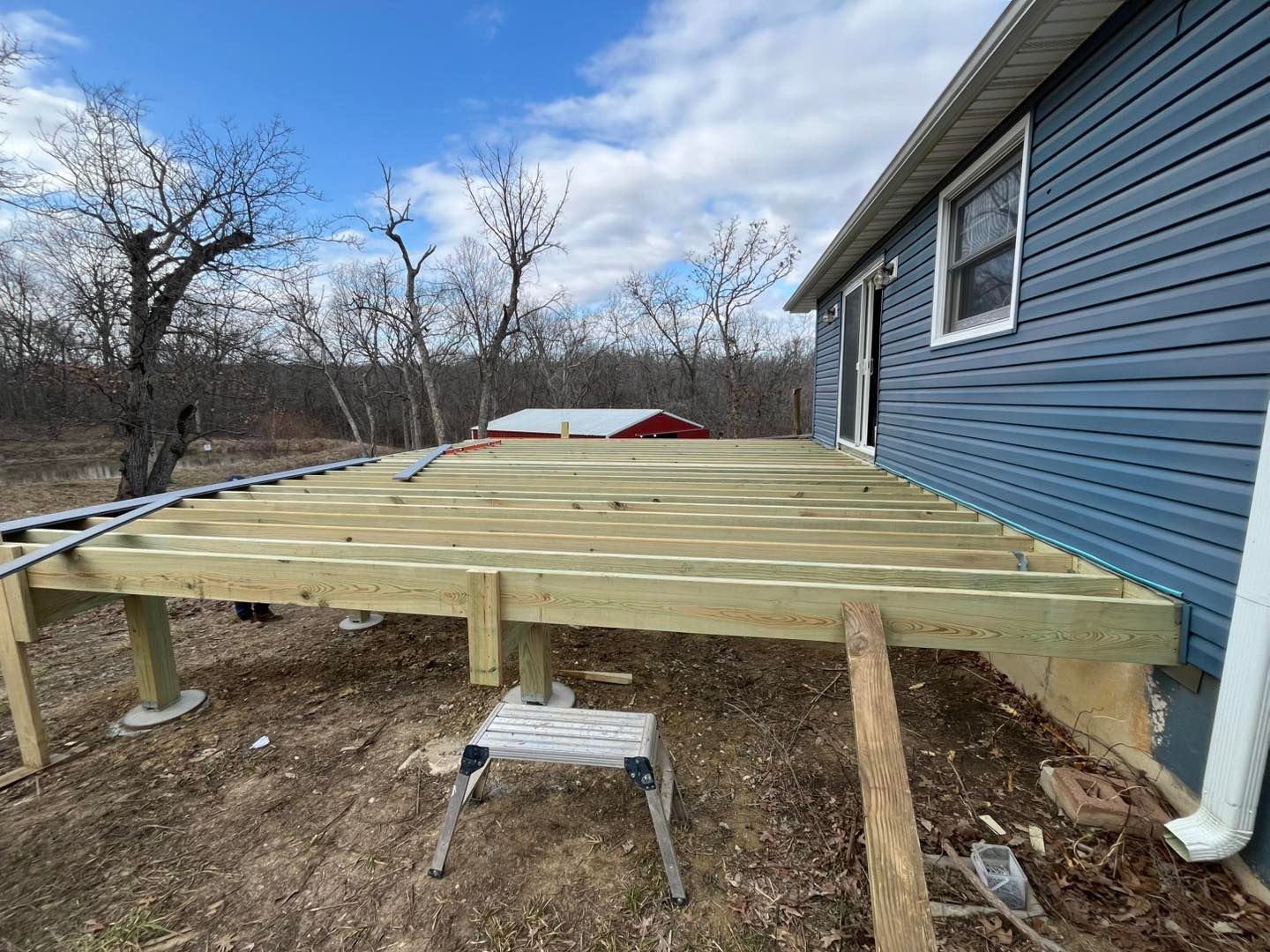 A partially constructed wooden deck next to a blue-sided house. The deck is supported by posts, with tools visible nearby.