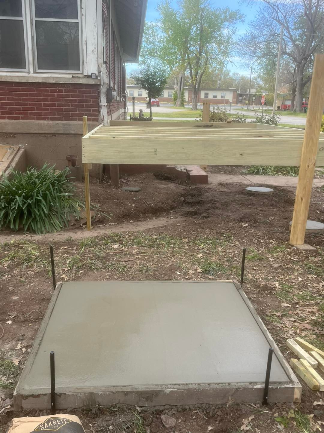Newly poured concrete pad in front of a house, with the beginning of a wooden deck framework visible in the background.