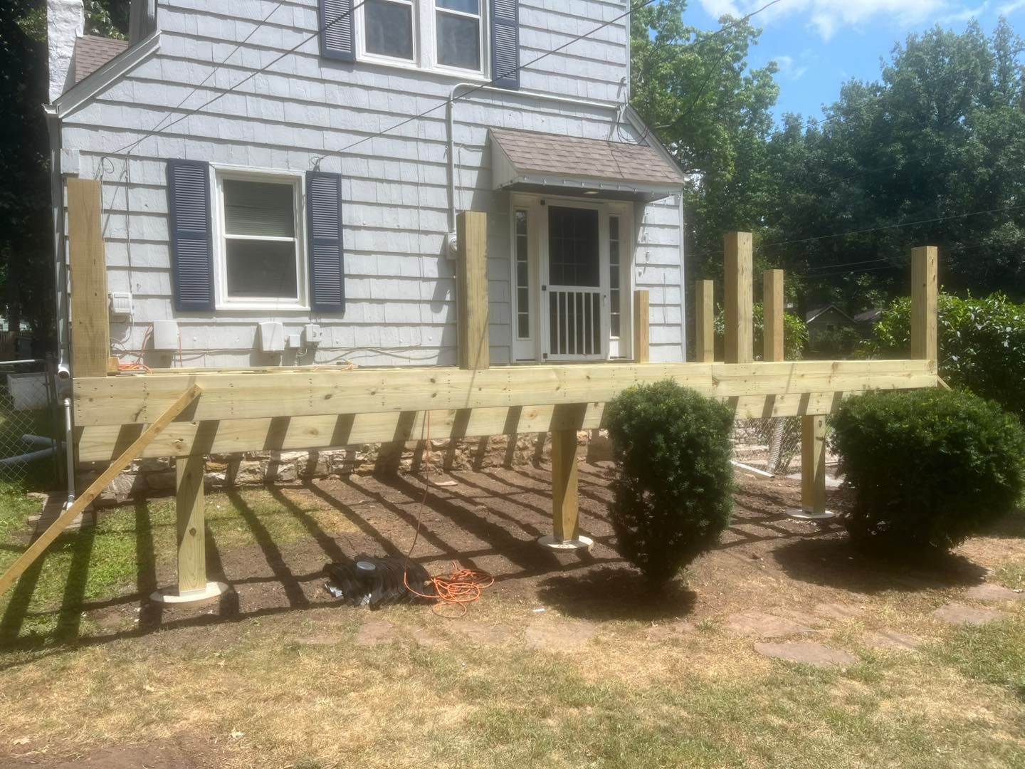 A partially built wooden deck in front of a light blue house with visible supports and railings. Green bushes are nearby.