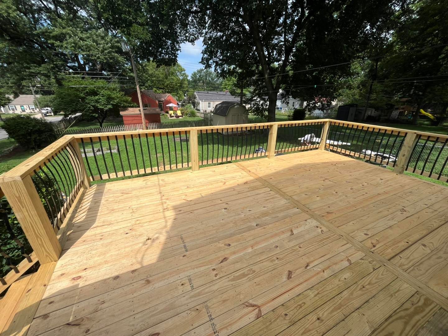 Wooden deck with black railing overlooking a green lawn, trees, and houses on a sunny day.