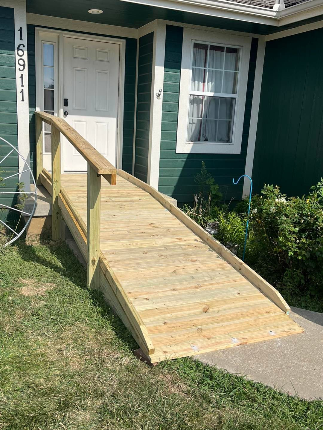 Wooden ramp leading up to the front door of a green house. The ramp has a handrail and rests on the grass.