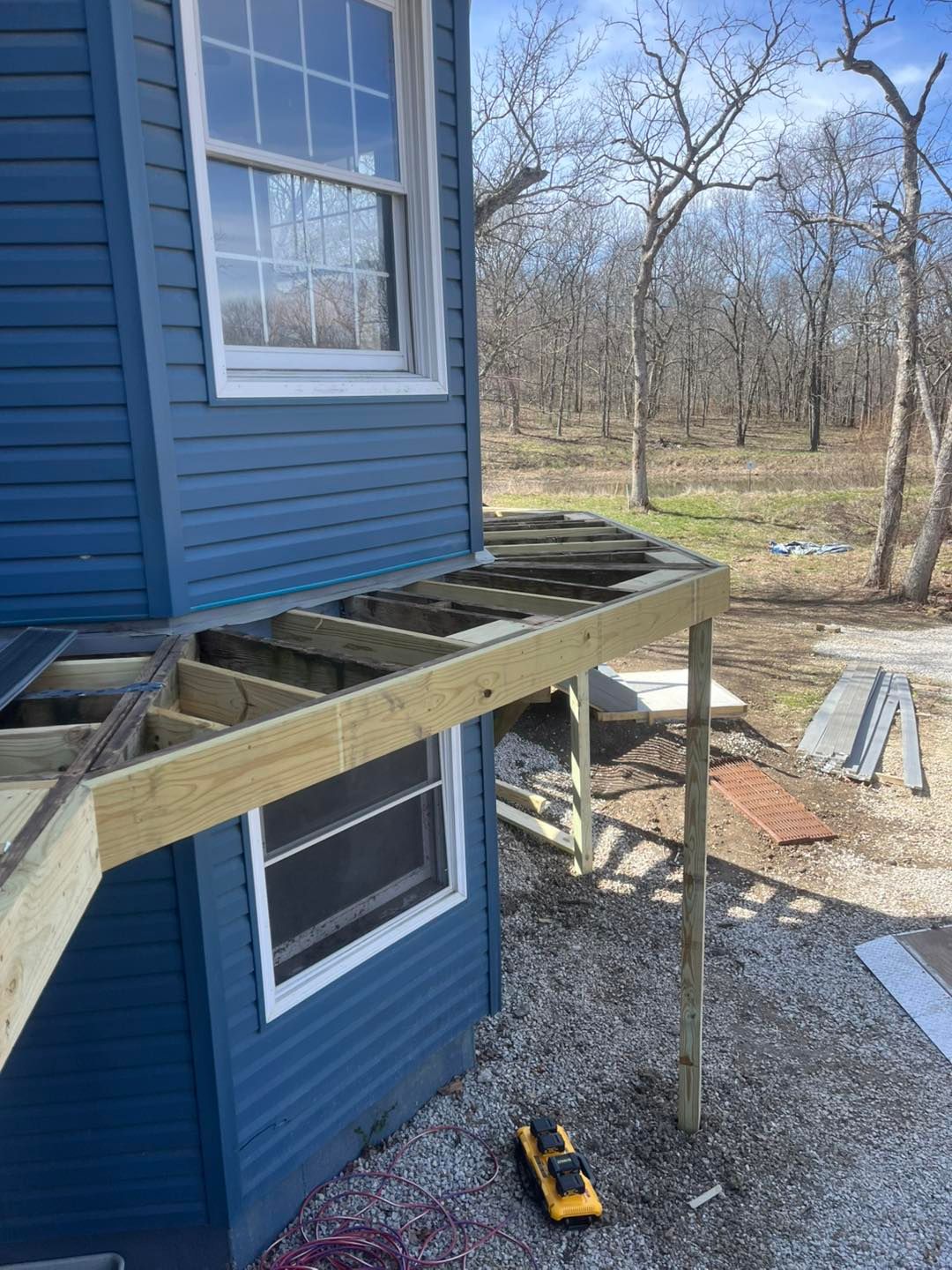 A deck under construction, attached to a blue house with a window. Wooden framing is visible, with trees in the background.
