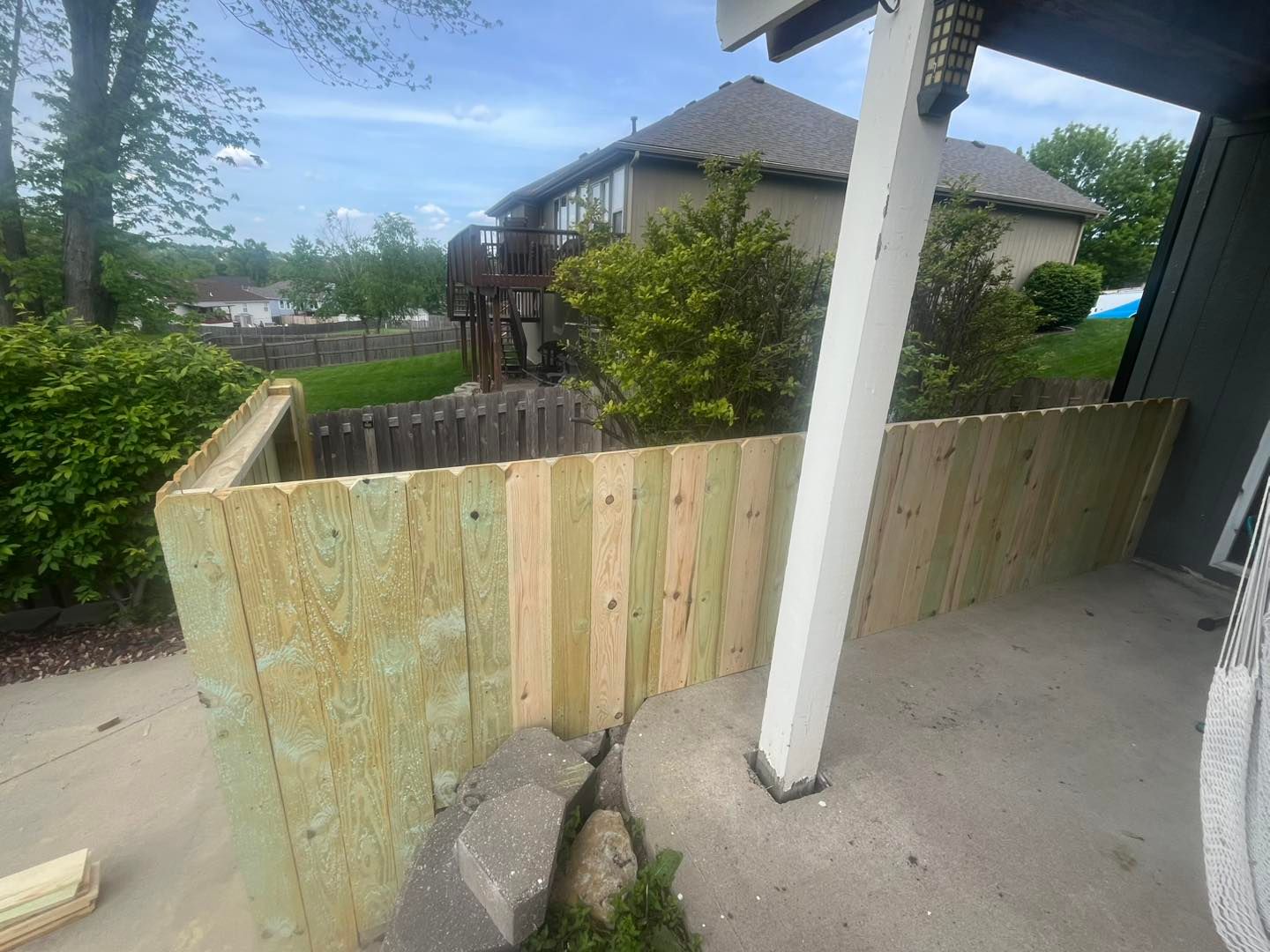 New wooden fence partially constructed along a patio, with a house and trees in the background.