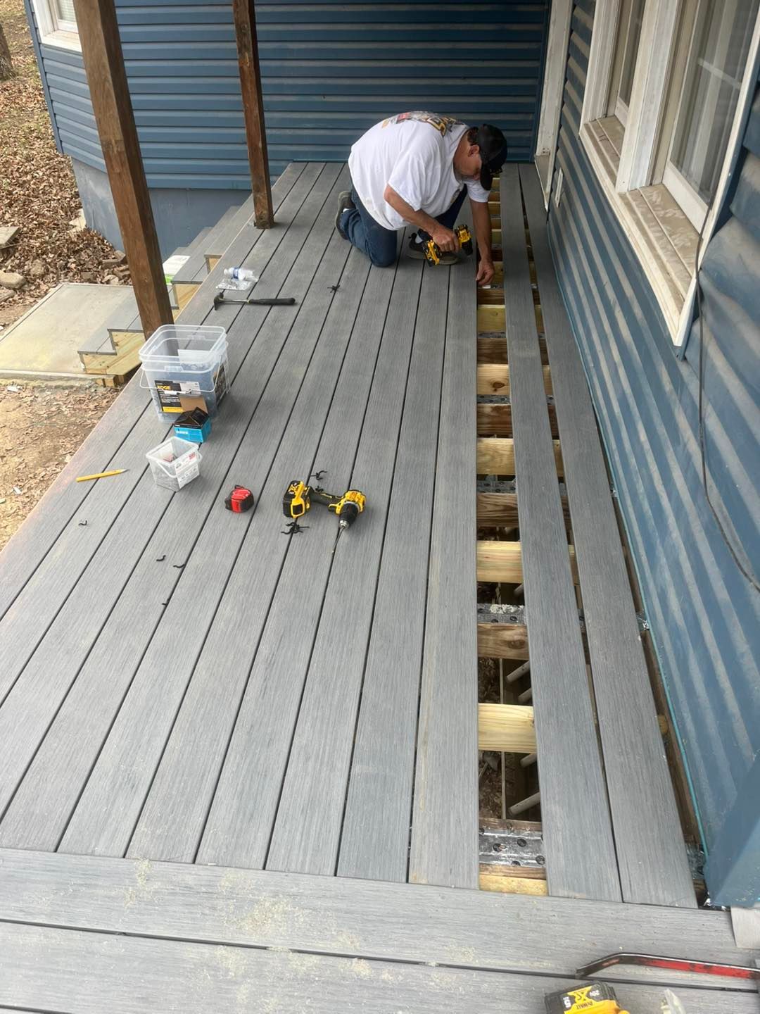 Man installing gray composite decking on a porch. He kneels, using a drill. The house exterior is blue.