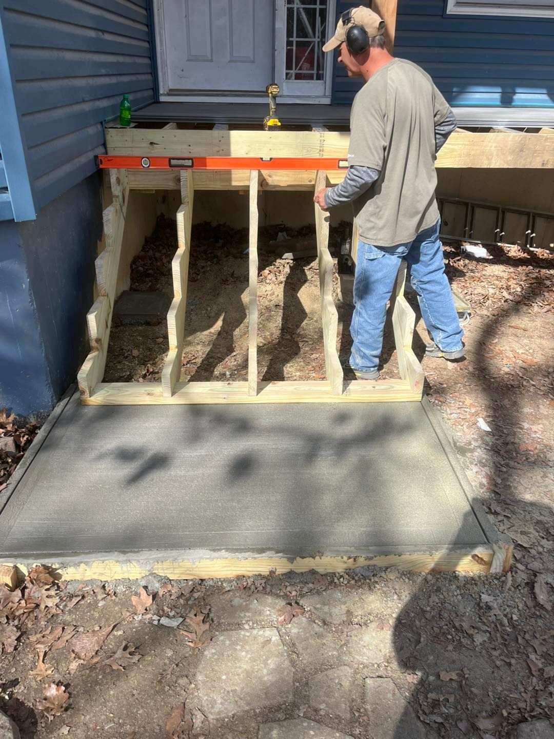 A man building a wooden stair frame next to a concrete slab, with a level in hand. Outdoors.