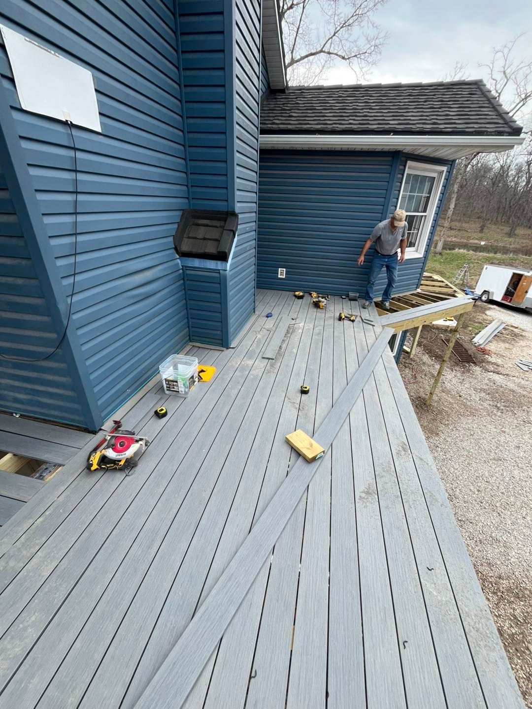A person works on a deck with gray composite planks. The blue house has a dark roof and is surrounded by gravel.