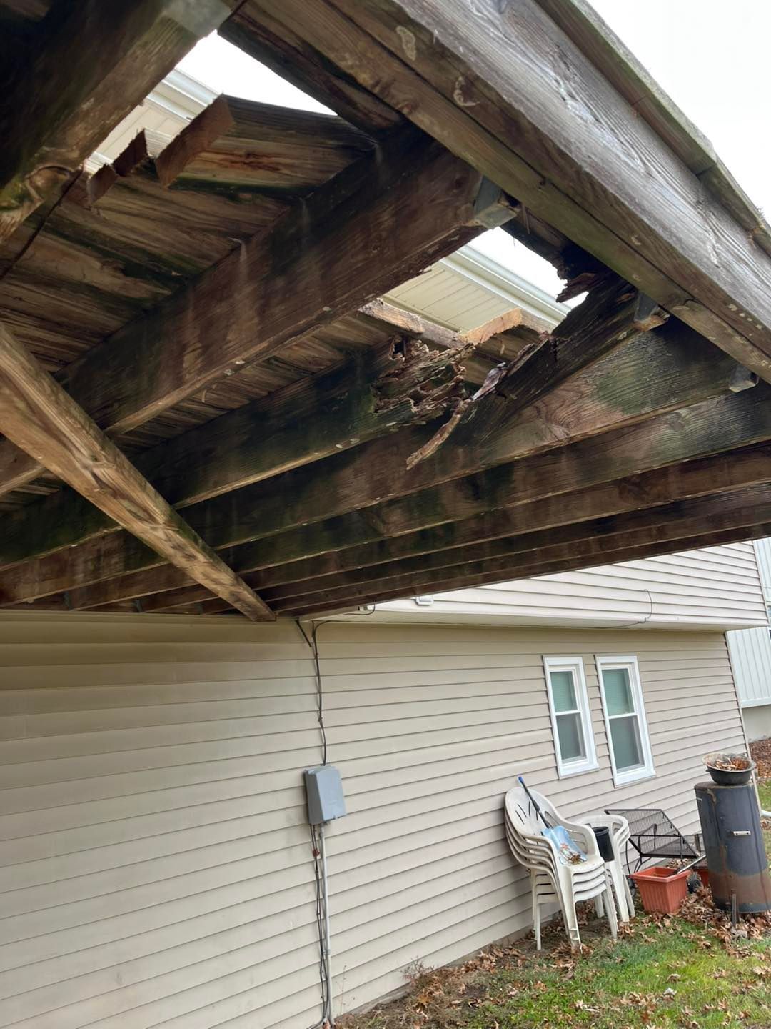 Damaged wooden porch overhang with a large hole; beige siding and a white house visible in the background.
