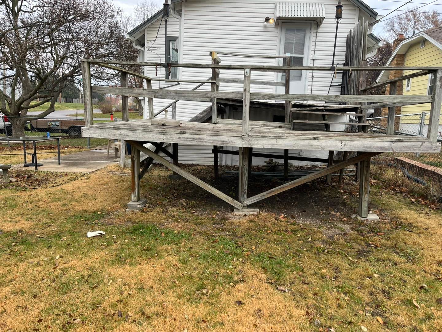 A weathered wooden deck attached to a white house, raised off the ground on support beams, in a yard with brown grass.