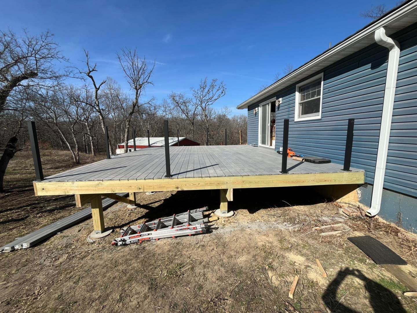 A deck constructed next to a blue house. It has black railings and grey decking. The setting is outdoors on a sunny day.