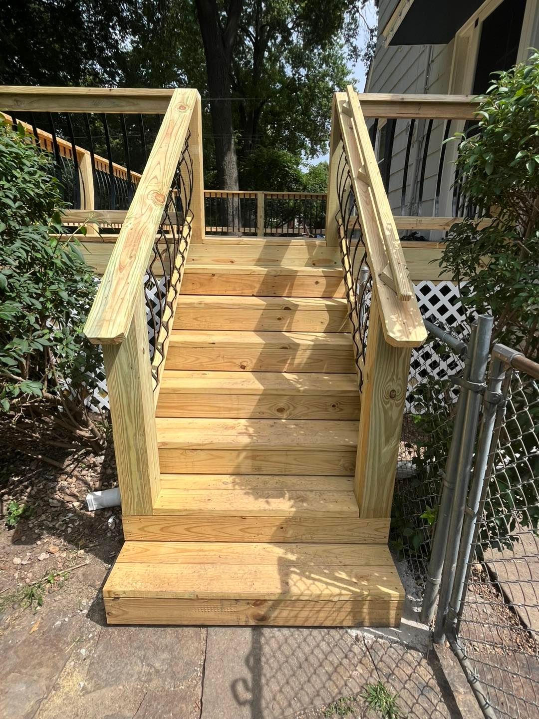 Wooden staircase leading up to a deck, flanked by railings with decorative black metal balusters. Outdoors on a sunny day.