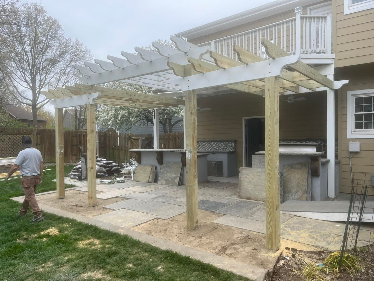 A partially constructed pergola attached to a house. A person walks near the structure. Light brown siding.