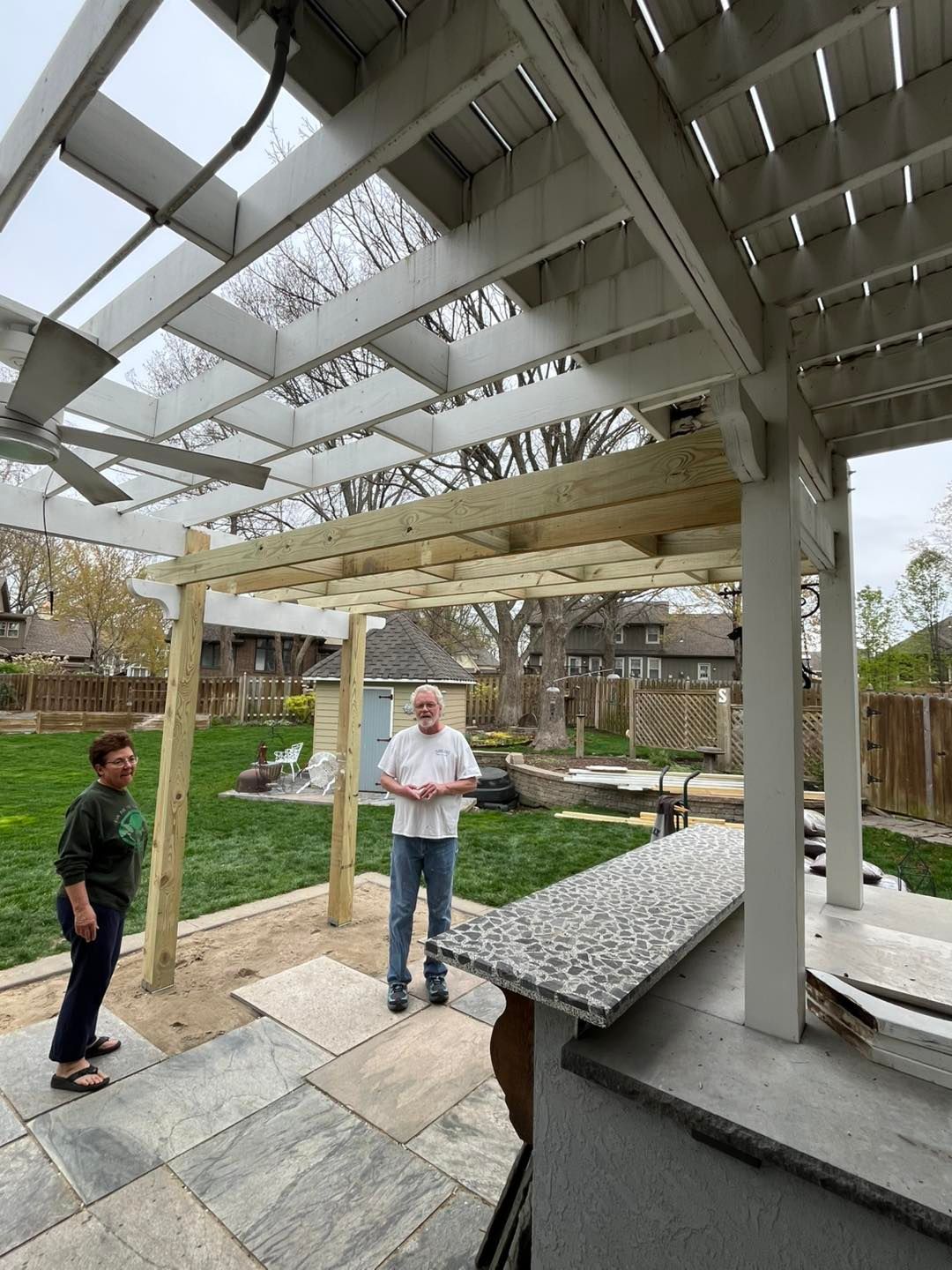 Two people standing under a pergola being built; one is woman, one is man, with a granite-topped bar in the foreground.