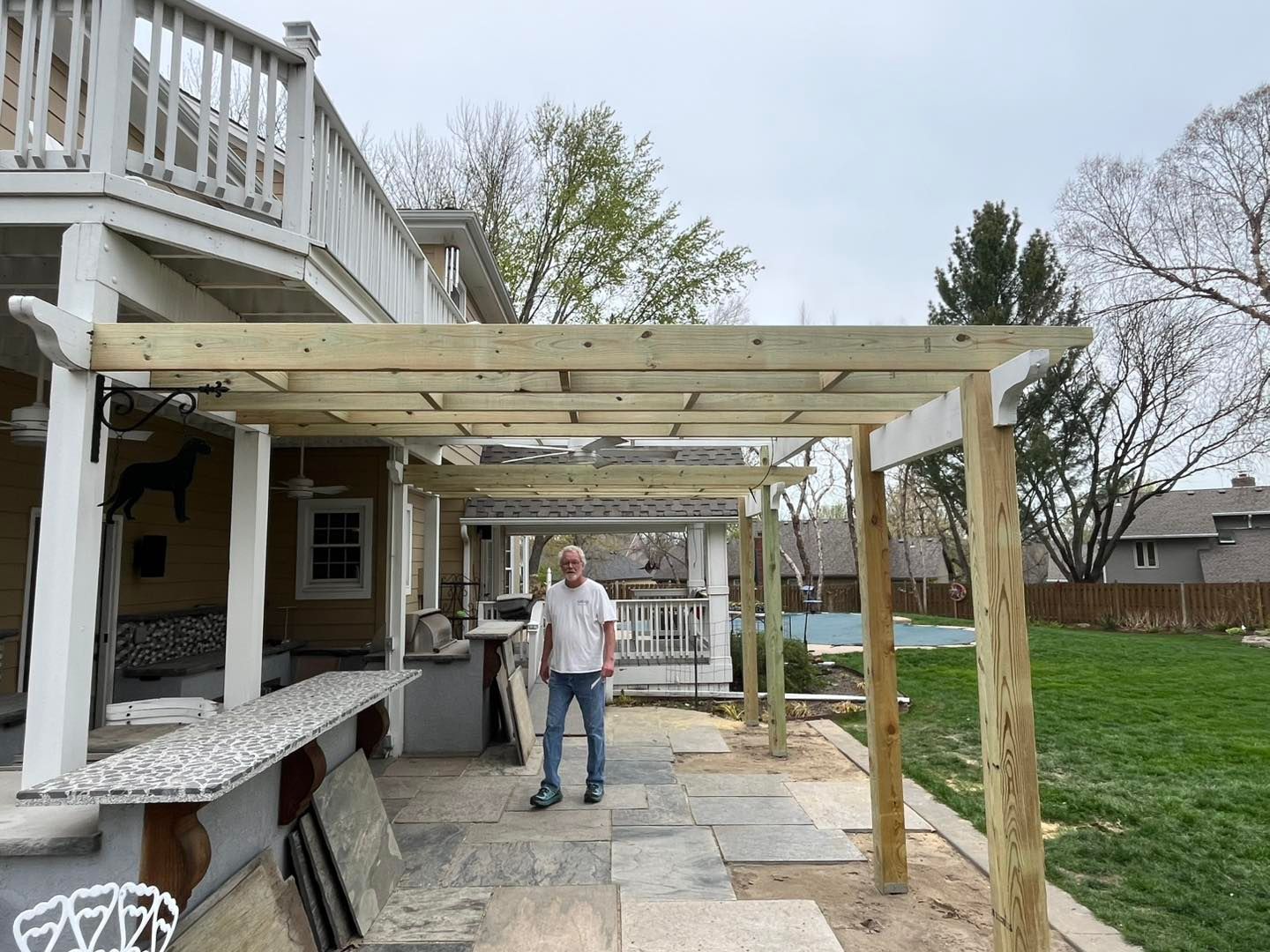 Man standing under a partially built wooden pergola attached to a house with a patio and backyard in view.