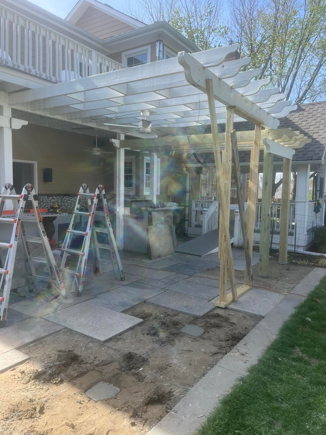 Construction of a wooden pergola on a patio. Ladders and materials are visible, with a house in the background.