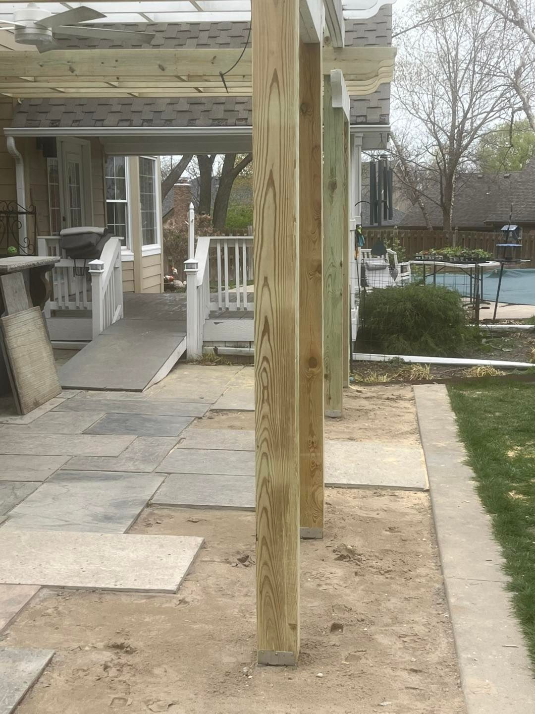 Wooden pergola posts in a dirt area next to a patio. A ramp leads to a white deck. Pool and lawn are in the background.