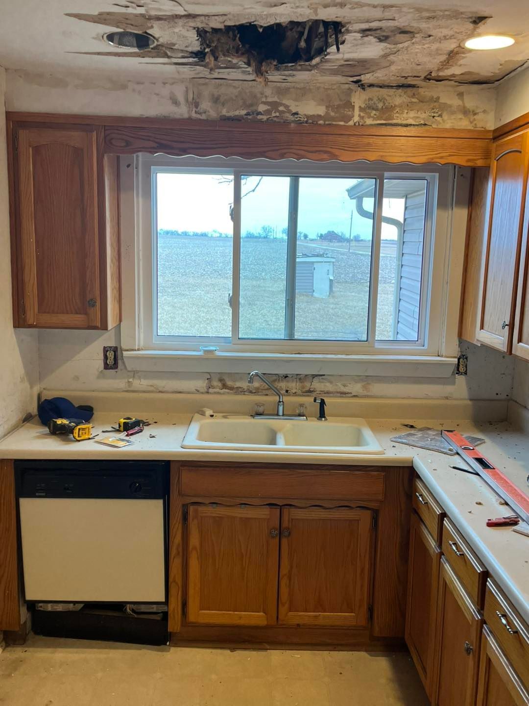 Kitchen with water damage on the ceiling above a sink. Cabinetry and window are visible, as well as tools on the countertop.