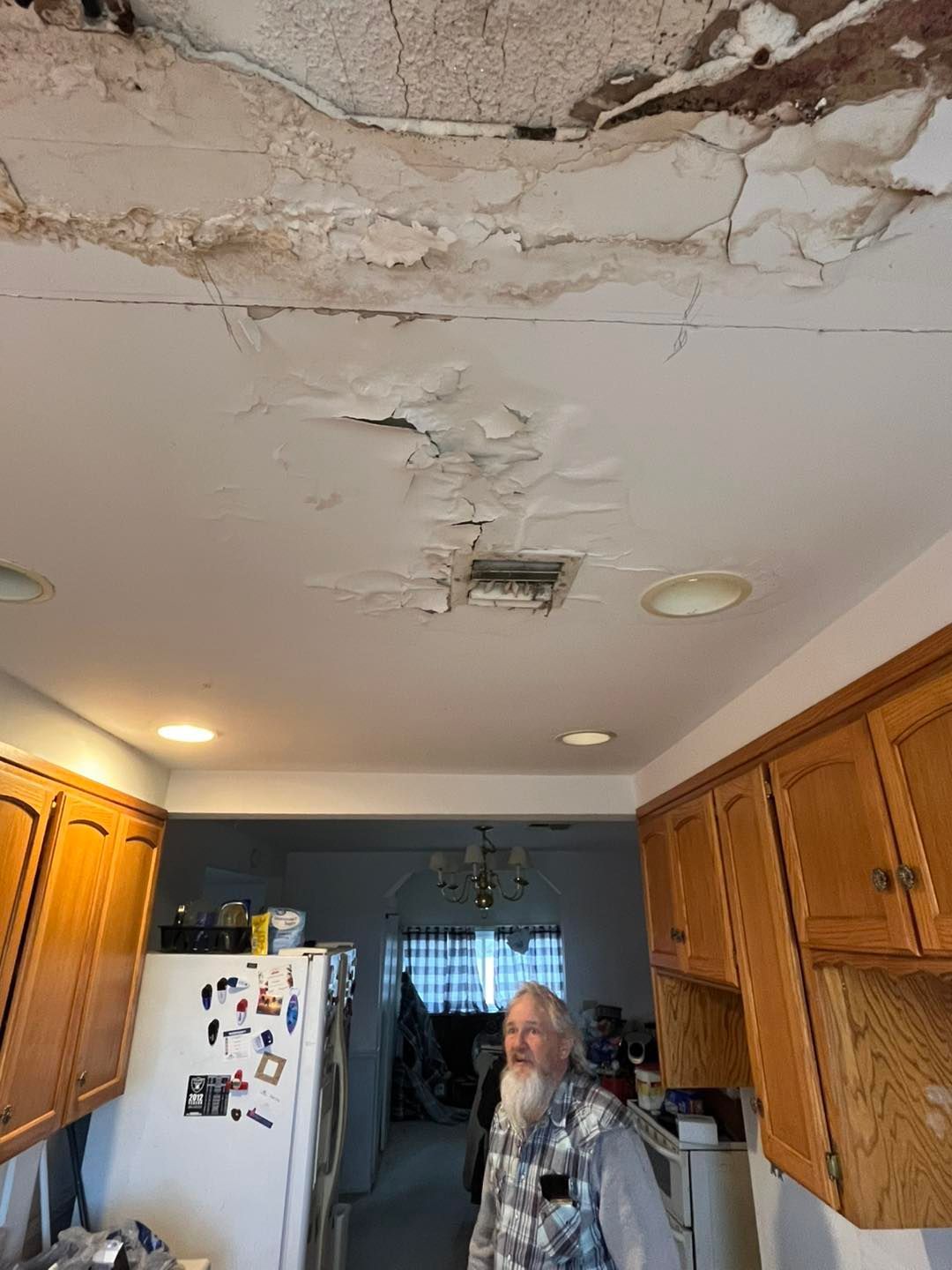 A man stands in a kitchen with a damaged ceiling showing water damage. Brown cabinets line the walls.