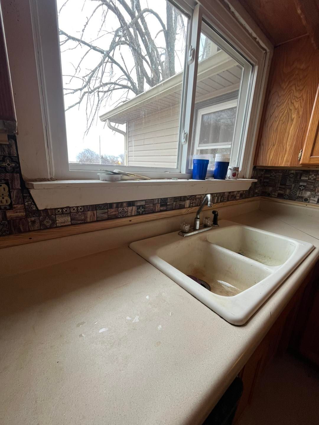 Kitchen sink under a window with a view of a house. Beige countertop and white sink with a faucet.