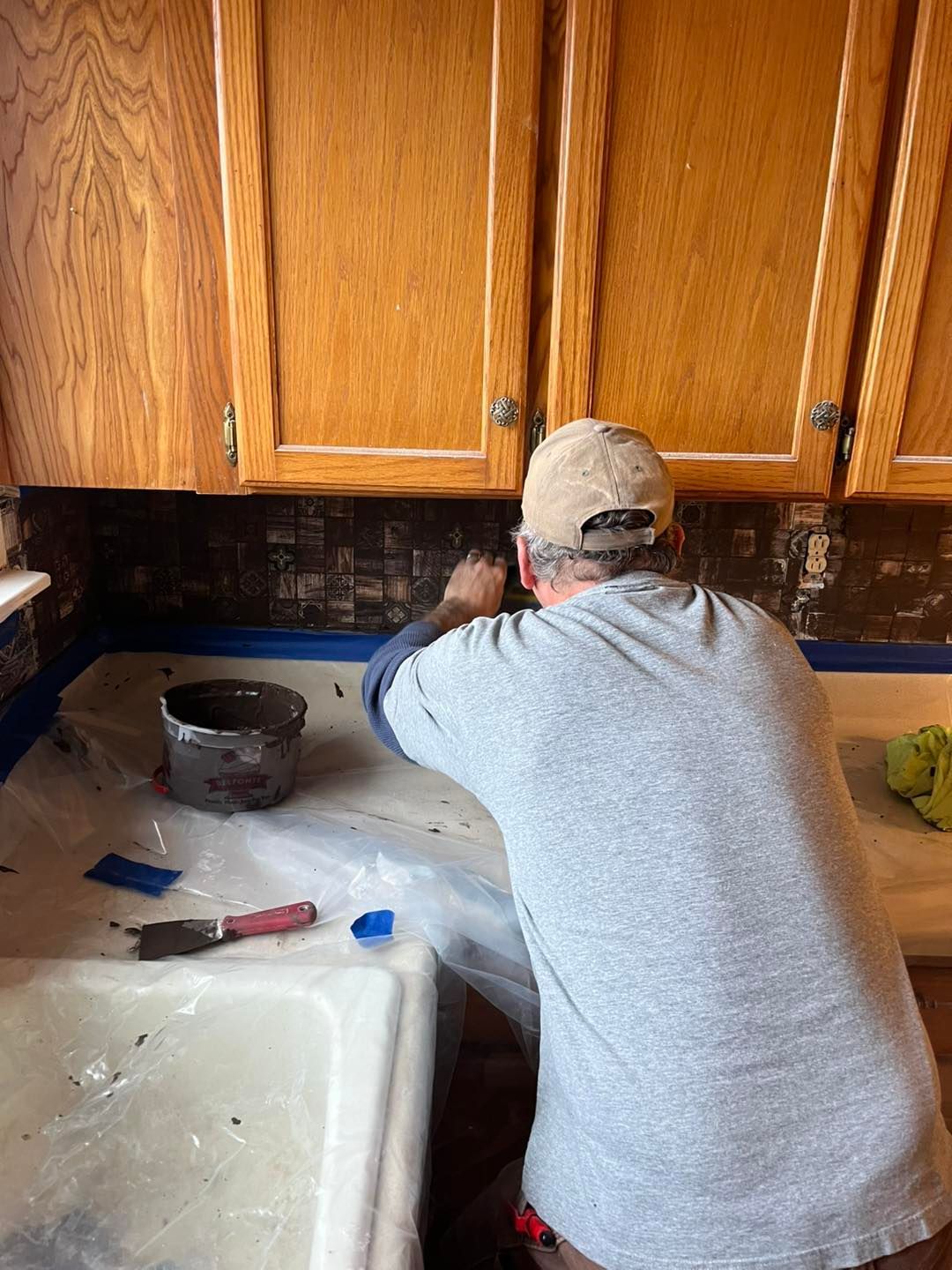 A person is tiling a kitchen backsplash. They're wearing a cap and gray shirt, 