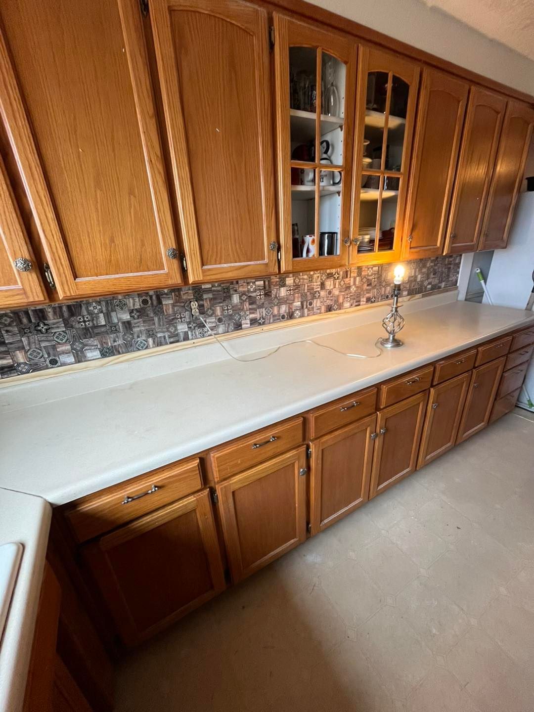 Kitchen with light-colored countertops, wooden cabinets, and a mosaic backsplash. A small lamp sits on the counter.