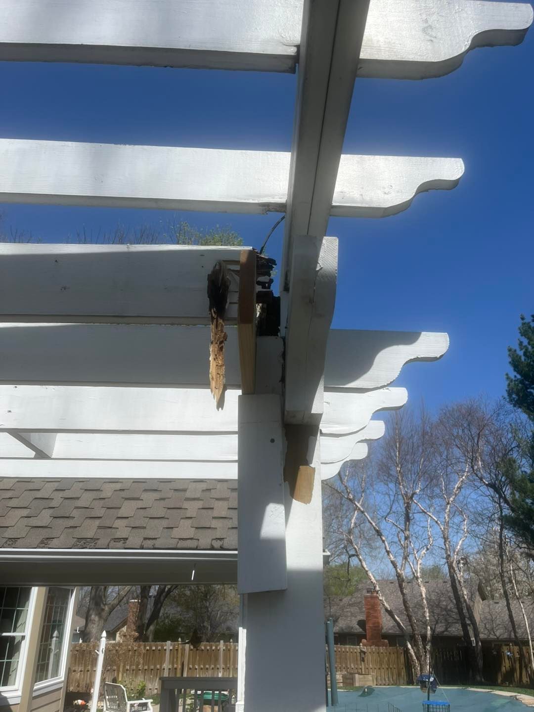 White pergola with damaged upper beam and support, set against a blue sky. The damage exposes wood and hardware.