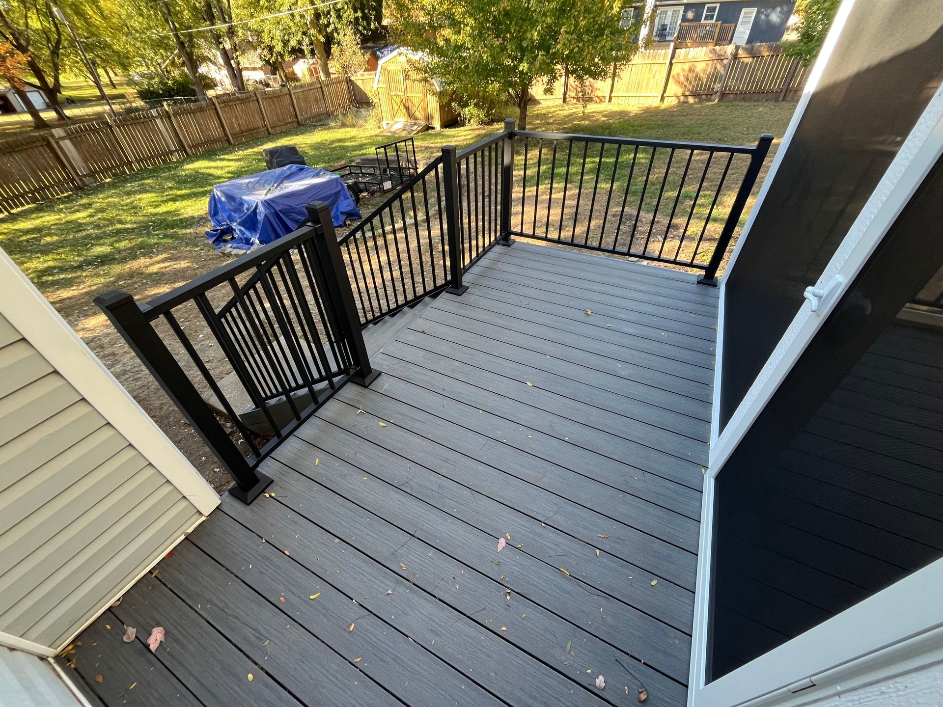 A composite deck with black railing and steps, leading to a backyard with a fence and covered furniture.