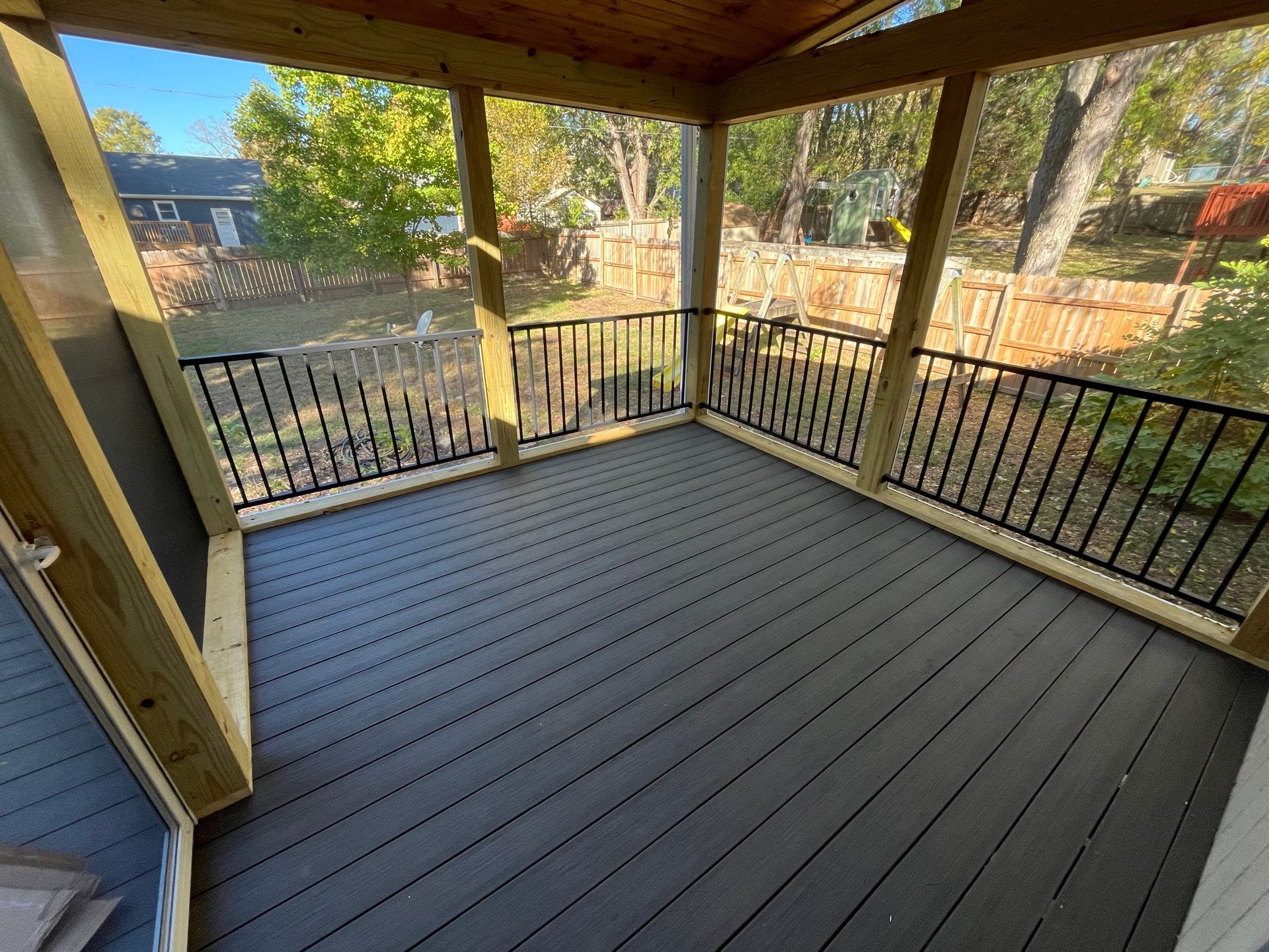 Screened-in porch with gray composite decking, black railing, and wooden posts, overlooking a backyard.