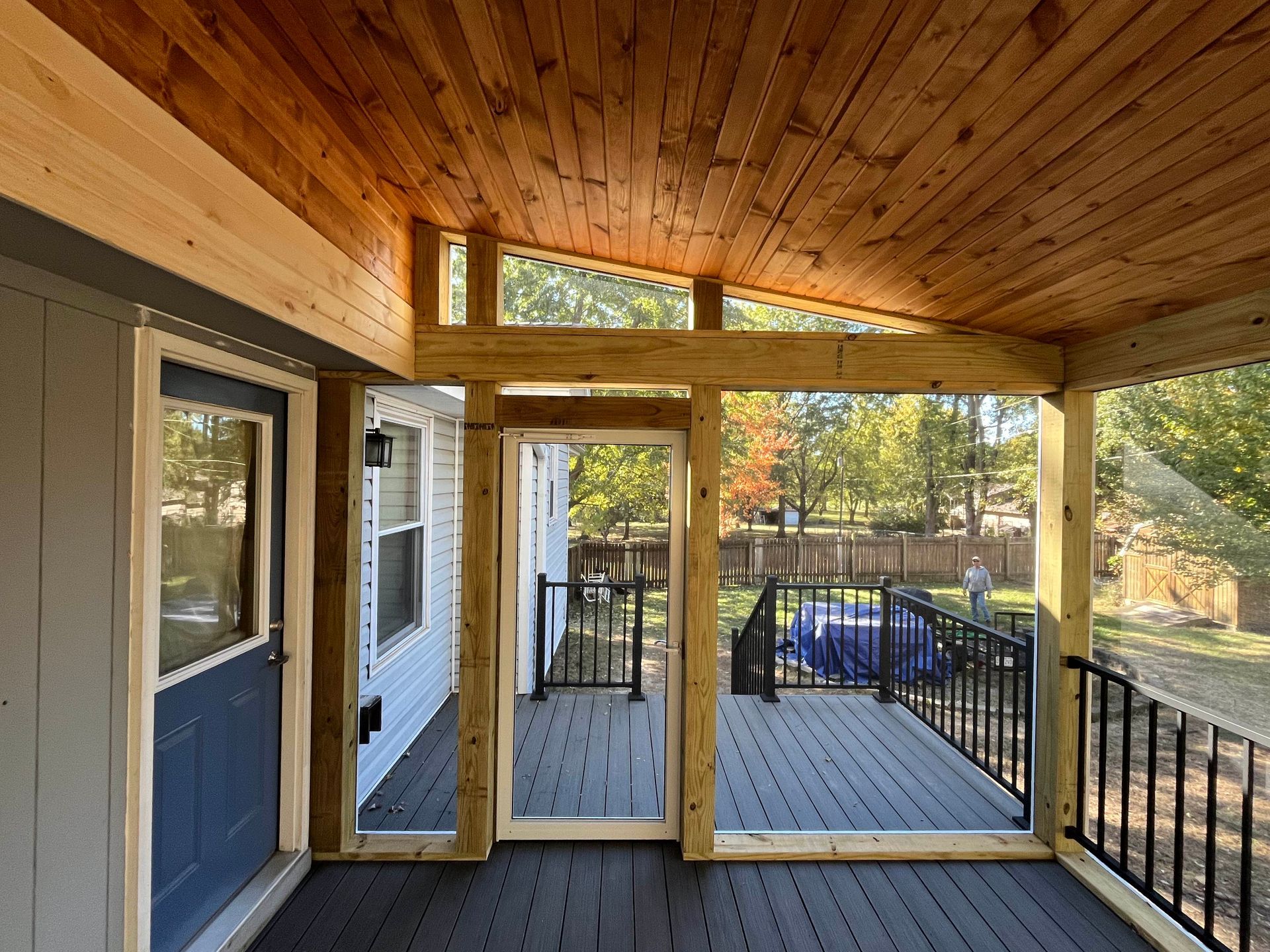 Covered wooden porch with black railing and door, overlooking a backyard with a fence.