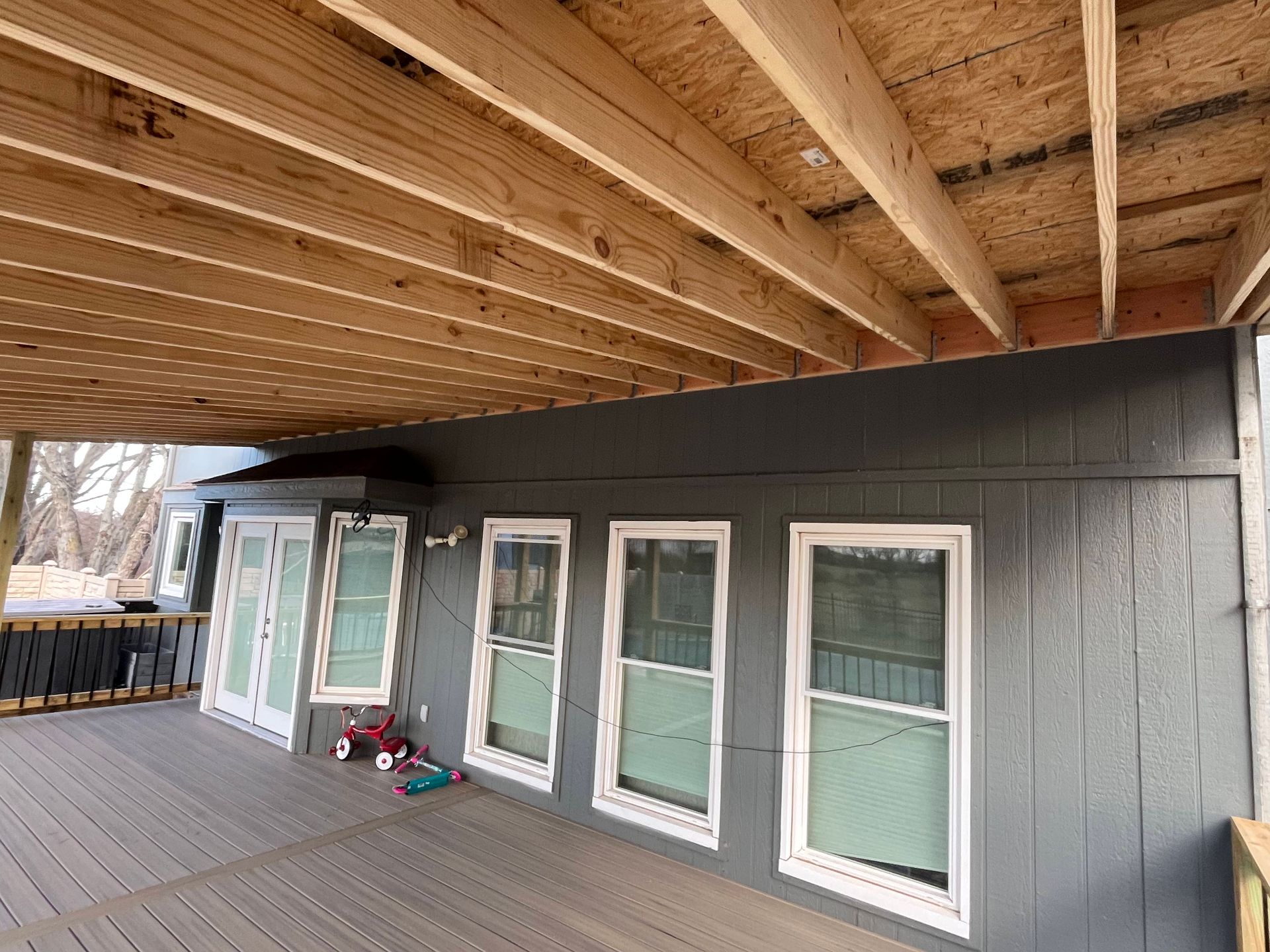Exterior view of a house with a deck. Gray siding, white-framed windows. Wooden beams under the deck.