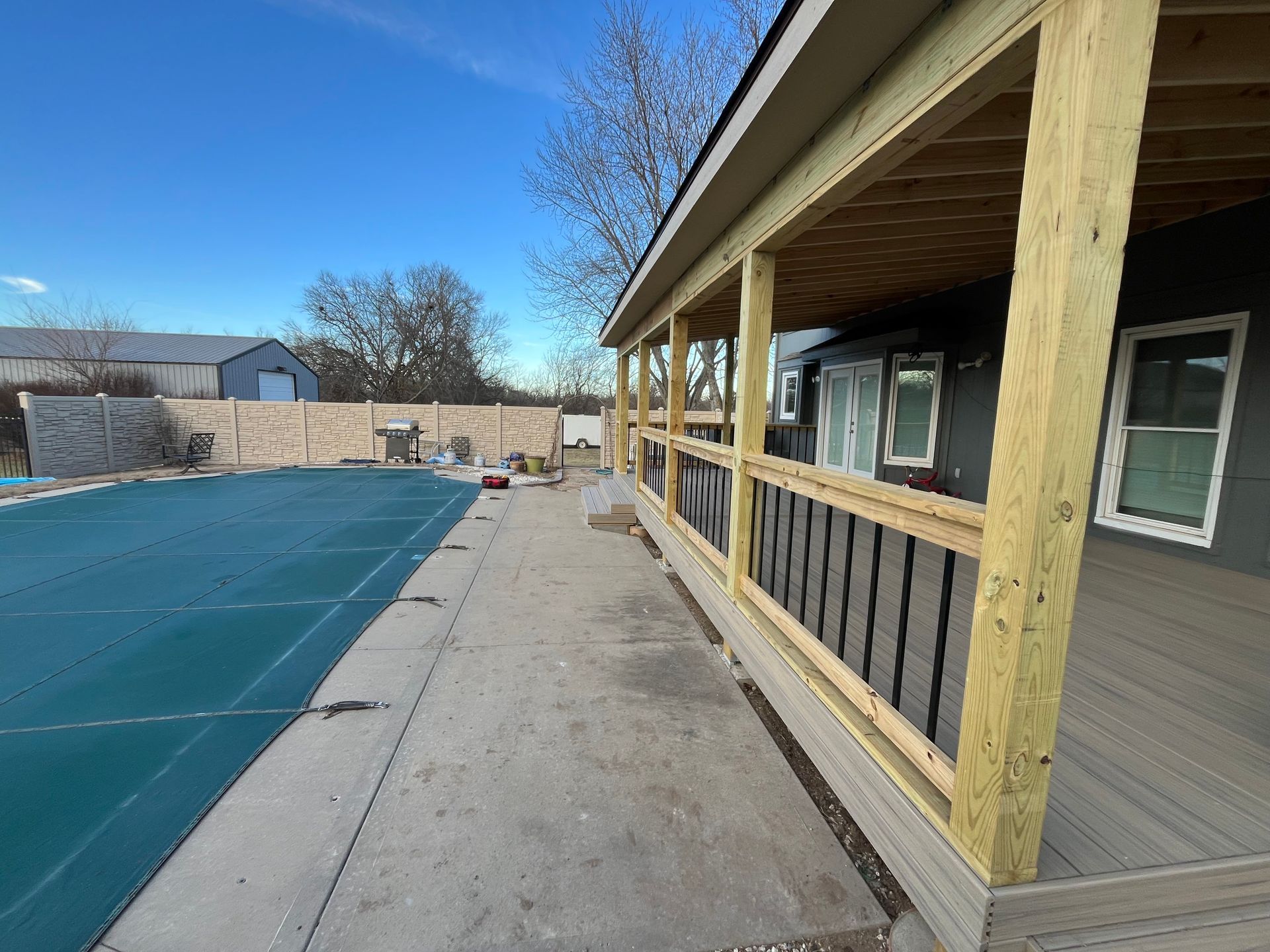 New wooden deck construction next to a pool. Building is gray with black railings. Clear blue sky.