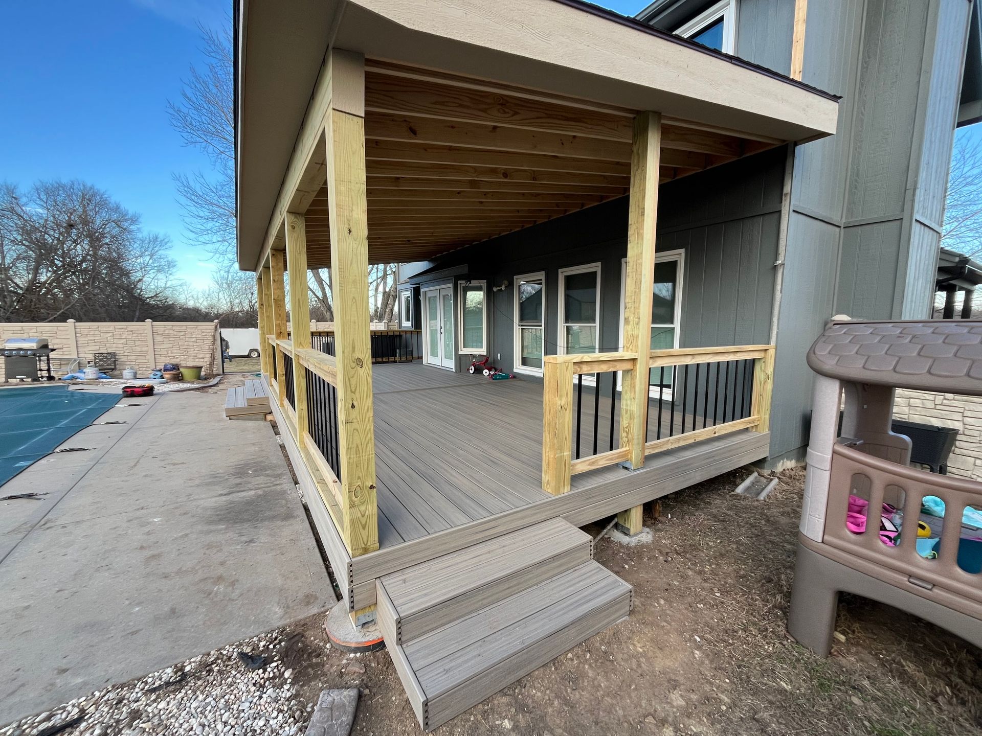Wooden deck with covered porch, steps, and black railing. Backyard setting, pool visible.