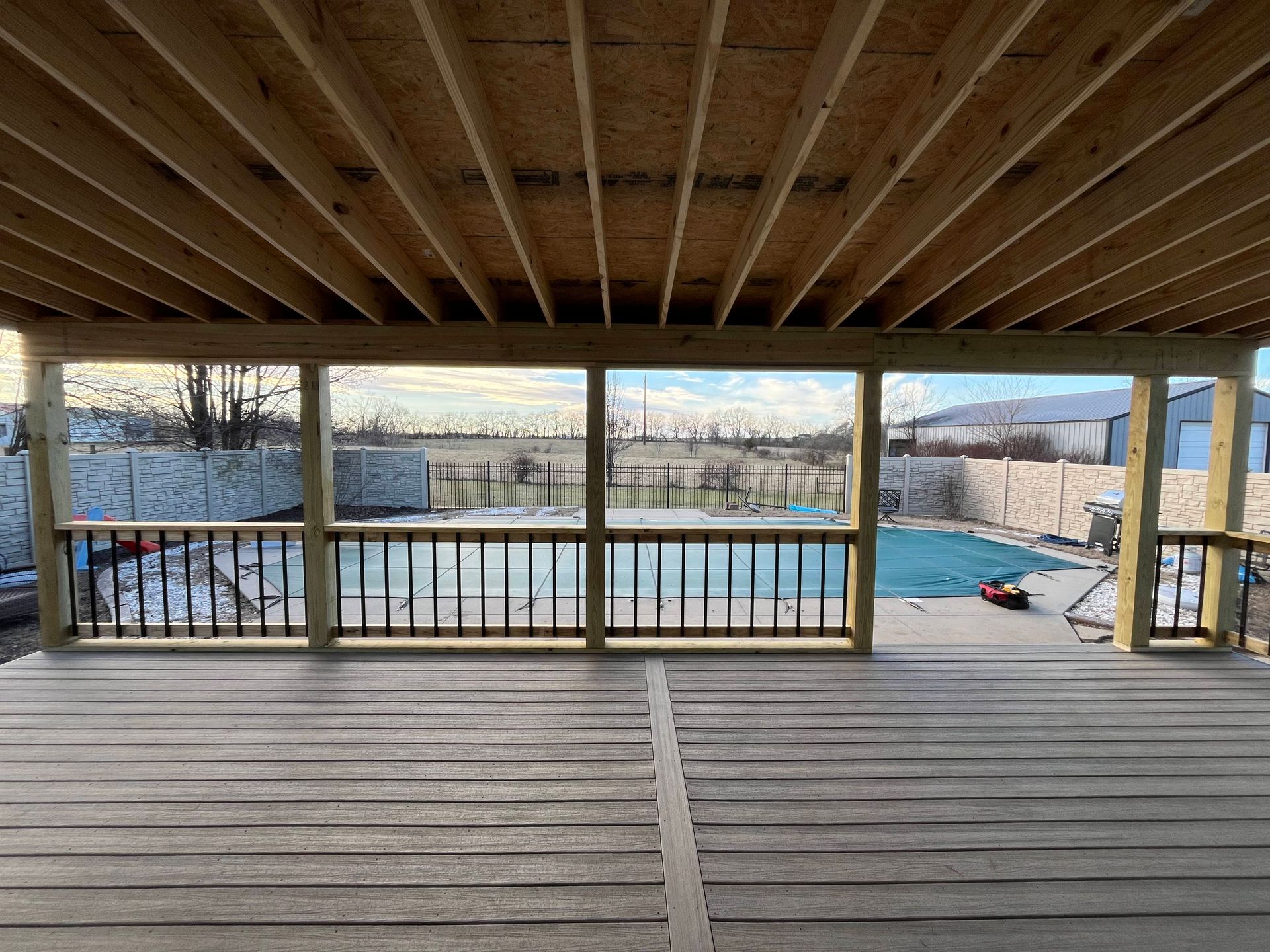 View from a covered deck overlooking a pool. Wooden construction. Grey decking and railings.