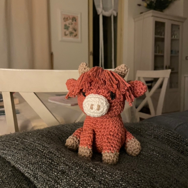 Cute Amigurumi Highland Cow Toy, chestnut brown with cream nose, beige feet and horns and lots of floppy hair.