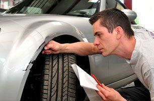 A man is looking at a tire on a car while holding a piece of paper.