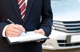 A man in a suit and tie is writing on a clipboard in front of a car.