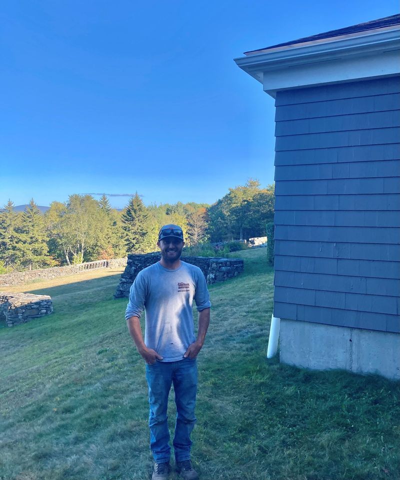 Man standing on a grassy hill, hands in pockets, next to a blue-sided building; trees and blue sky background.