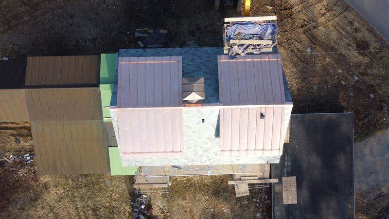 Aerial view of a building under construction, featuring copper roofing, asphalt shingles, and surrounding structures on a brown surface.