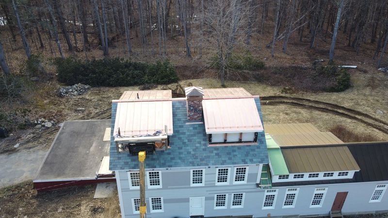 Overhead view of a light gray building with multiple roof sections of varying colors, including a brick chimney. Trees in background.