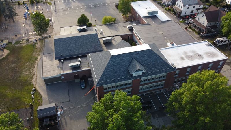 Aerial view of a brick building with a dark gray roof, surrounded by trees and a paved area.
