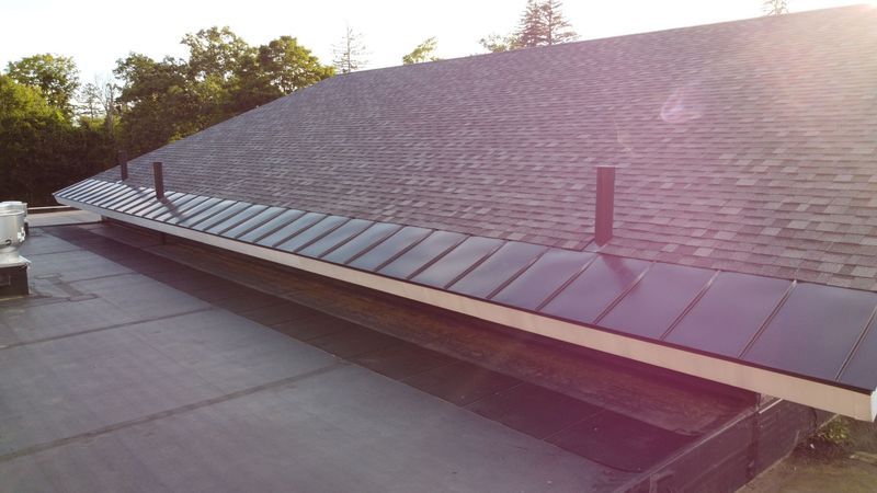 Black flat roof with gutter, metal panels, and a sloped, shingled roof in the background.