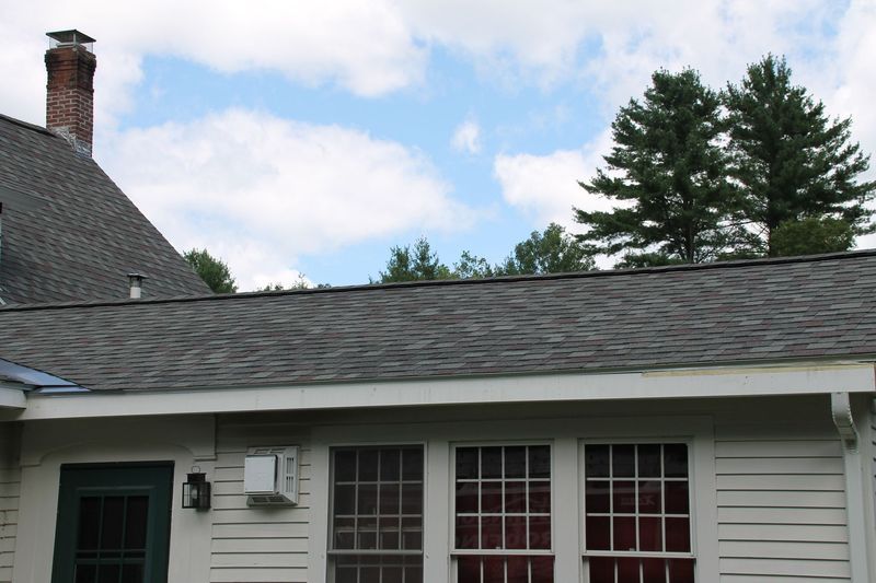Shingled roof with a chimney and part of a white building with windows under a blue sky.