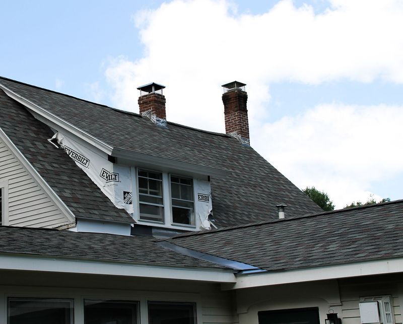 House roof with chimneys, some sections covered in roofing material, others showing underlayment, and windows.