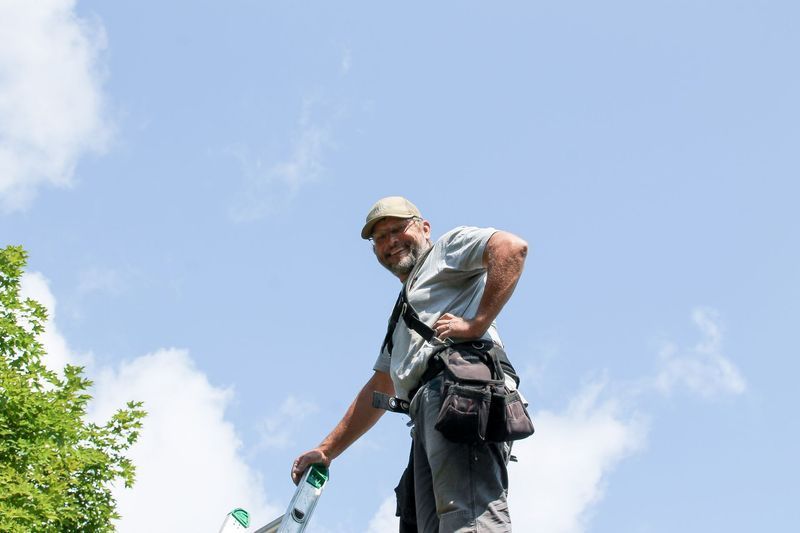 Man on a ladder smiling, wearing a tool belt, looking up at the sky.