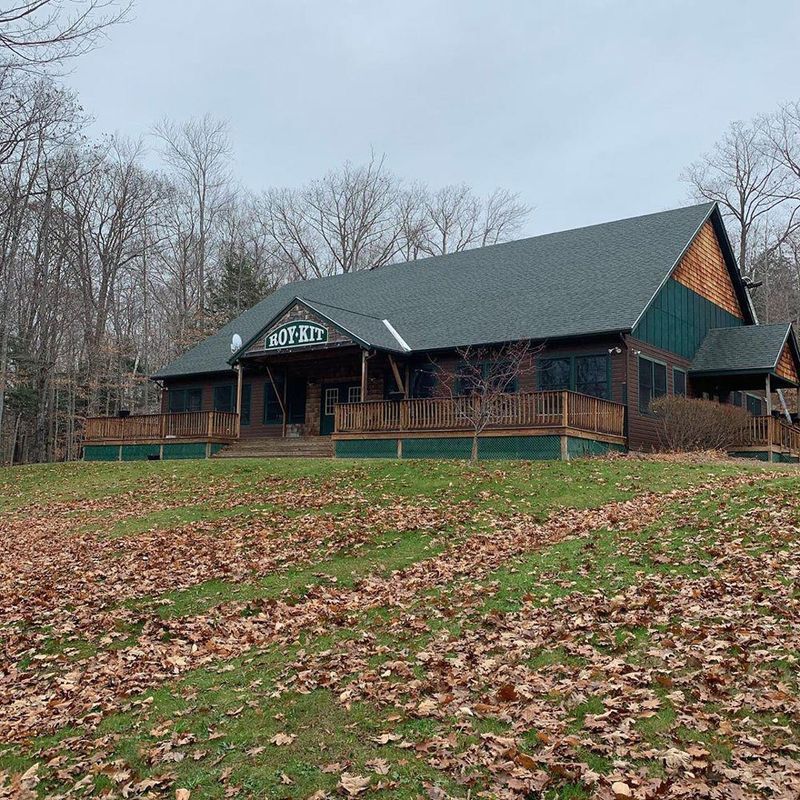 Brown building with a dark green roof and wooden deck, in a wooded area with a grassy hill covered in fall leaves. Brown building with a dark green roof and wooden deck, in a wooded area with a grassy hill covered in fall leaves.