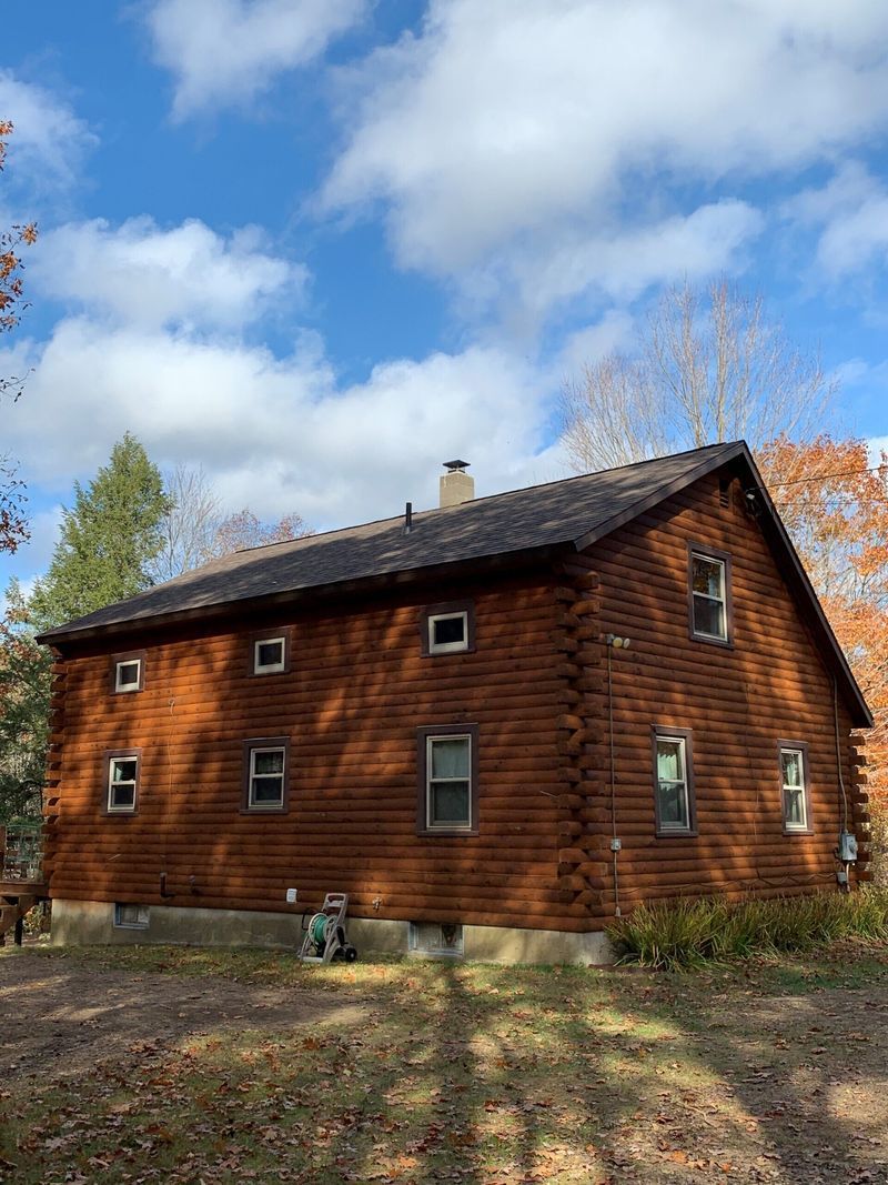Log cabin house with brown logs, small windows, and a dark roof under a blue sky with clouds.