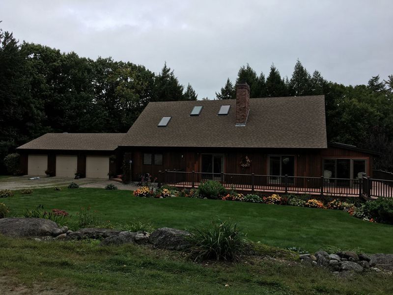 Brown house with garage and deck, surrounded by green grass and trees. Overcast sky.