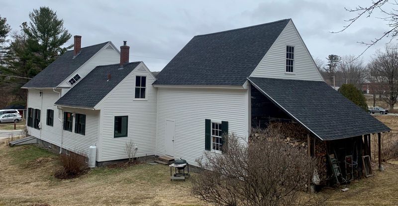 White two-story house with black shingled roofs and green-trimmed windows under a cloudy sky.